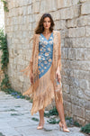 Woman wearing a beige lace coverup with fringe over a blue floral dress, standing against a stone wall.
