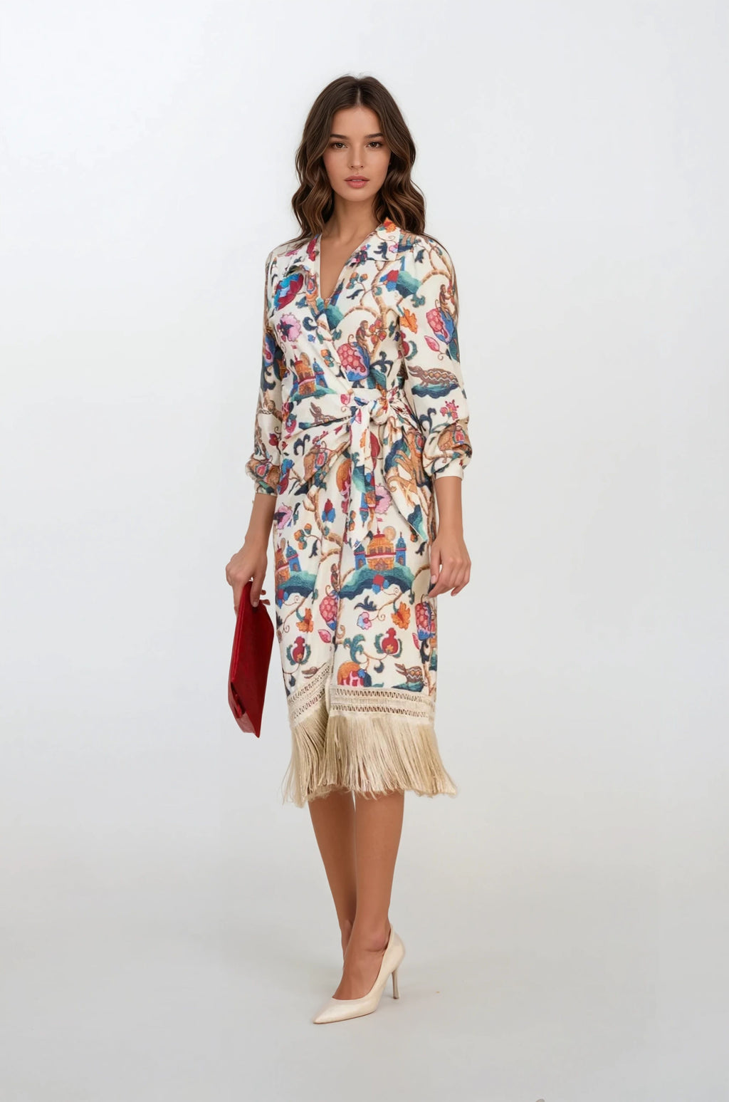 Woman wearing a cream wrap dress with multicolored cultural print and fringe hem, holding a red clutch and styled with white heels.