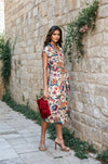 Model wearing a sleeveless printed wrap dress with elephant and floral motifs, styled with nude heels and a red clutch, standing against a stone wall.