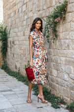 Model wearing a sleeveless printed wrap dress with elephant and floral motifs, styled with nude heels and a red clutch, standing against a stone wall.