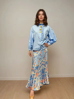 Woman wearing a sky blue cotton blouse with balloon sleeves, front ruffle trim, and yellow floral appliqués, paired with a flowy floral maxi skirt and nude heels, standing against a plain neutral wall