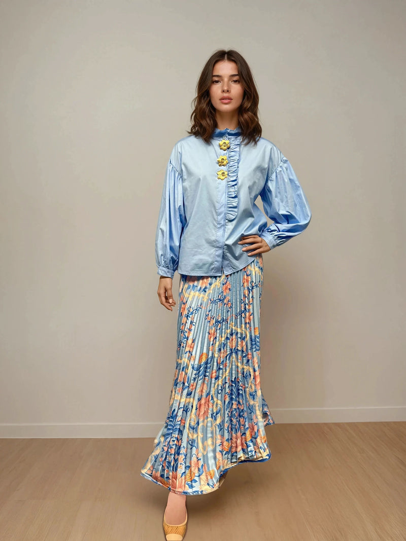 Woman wearing a sky blue cotton blouse with balloon sleeves, front ruffle trim, and yellow floral appliqués, paired with a flowy floral maxi skirt and nude heels, standing against a plain neutral wall