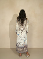 Back view of woman wearing long boho-inspired maxi dress with sun and moon design, blue butterflies, and script accents, styled with heeled boots.