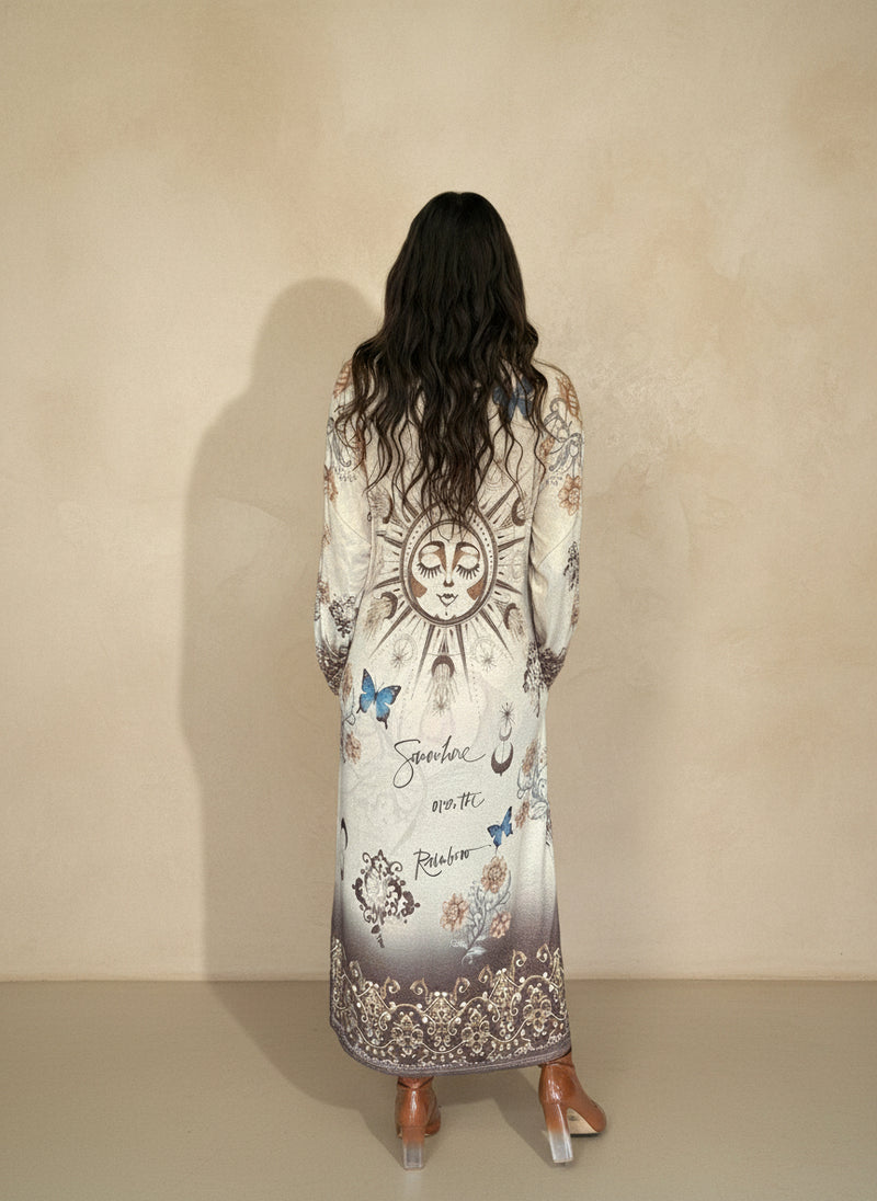 Back view of woman wearing long boho-inspired maxi dress with sun and moon design, blue butterflies, and script accents, styled with heeled boots.