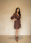 Model wearing a brown polka-dot ruffle mini dress with long puff sleeves, V-neckline, and waist belt, paired with brown ankle boots against a beige wall.