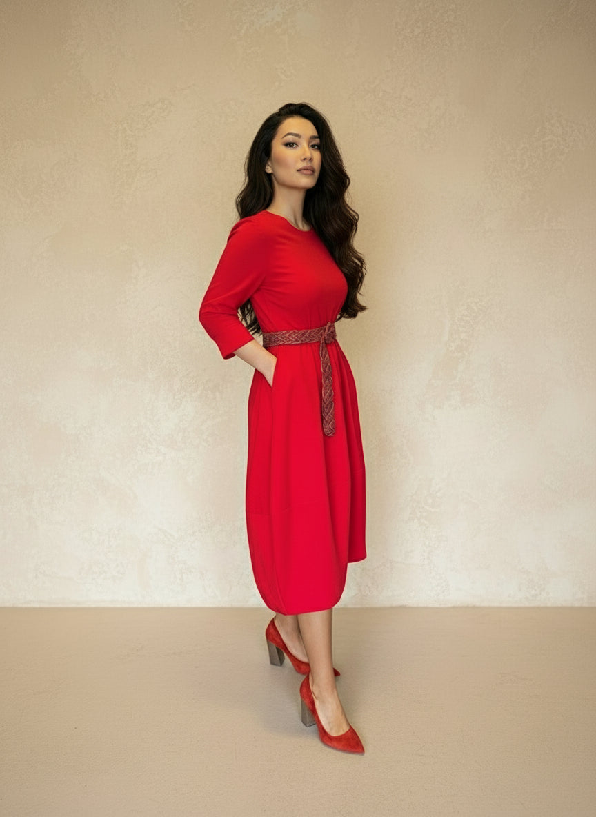 Model wearing a vibrant red midi dress with three-quarter sleeves and a braided waist belt, paired with red heels, standing against a neutral beige wall.