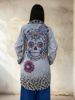 Back view of a woman wearing a long gray jacket with a colorful embroidered sugar skull and floral design, paired with wide-leg blue jeans, standing against a textured stucco wall.