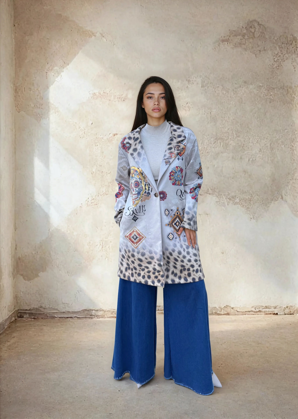 Front view of a woman in a patterned long gray jacket with sugar skull and floral embroidery, layered over a light gray turtleneck and wide blue trousers, posing indoors against a rustic wall.