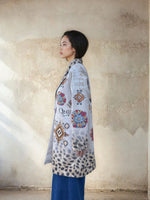 Side profile of a woman modeling a gray printed long coat featuring vibrant Aztec-inspired motifs and animal print gradient hem, styled with blue wide-leg denim pants.