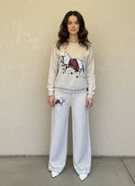 Woman wearing a cream knit lounge set with a white horse and red saddle motif, styled with white pointed heels.