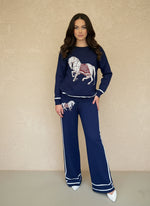Woman wearing a navy knit lounge set featuring a white horse graphic on the sweater and pants, styled with white heels.
