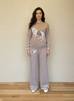 Woman modeling a taupe knit matching sweater and pants set decorated with horse embroidery and white accents.