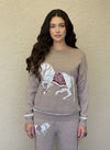 Close-up of a woman wearing a taupe knit sweater and pants with a white horse and burgundy saddle design.