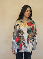 Model wearing a satin button-up blouse featuring bold leopard print, red and cream floral patterns, and a geometric black-and-white checkered design, paired with dark jeans.