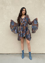 Woman modeling a short brown dress with blue floral print, ruffled neckline, and flared sleeves, paired with denim blue ankle boots, standing against a beige textured wall.