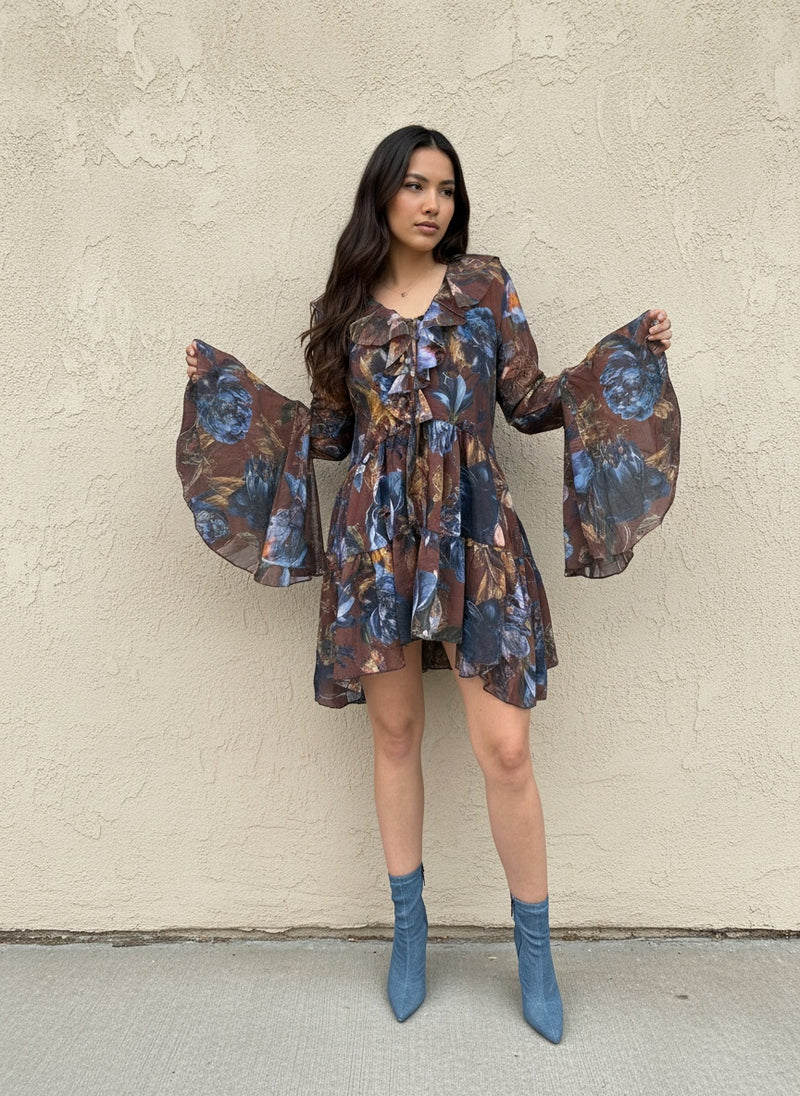 Woman modeling a short brown dress with blue floral print, ruffled neckline, and flared sleeves, paired with denim blue ankle boots, standing against a beige textured wall.
