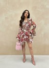 Fashion model in a pink floral ruffle mini dress with tiered skirt and flared sleeves, accessorized with a fluffy pink handbag and blush ankle boots, posing outdoors.