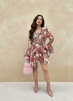 Fashion model in a pink floral ruffle mini dress with tiered skirt and flared sleeves, accessorized with a fluffy pink handbag and blush ankle boots, posing outdoors.