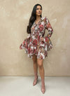 Model wearing a short floral ruffle dress with pink and beige tones, featuring long bell sleeves and layered hem, styled with blush pink ankle boots against a neutral background.
