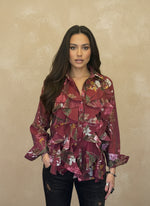 Woman wearing a burgundy floral chiffon blouse with cascading ruffles, long sleeves, and button-down front, posing against a beige background.