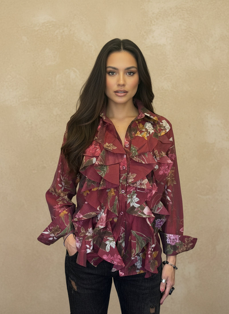 Woman wearing a burgundy floral chiffon blouse with cascading ruffles, long sleeves, and button-down front, posing against a beige background.