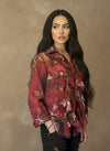 Woman wearing a burgundy floral ruffled blouse with long sleeves, posing against a tan wall. The blouse features layered ruffles and a sheer, elegant texture.