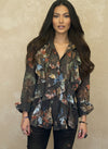 Woman in a dark brown floral ruffled blouse with blue, orange, and green floral print, styled with dark jeans and minimal accessories, posing confidently against a beige wall.