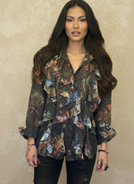 Woman in a dark brown floral ruffled blouse with blue, orange, and green floral print, styled with dark jeans and minimal accessories, posing confidently against a beige wall.