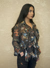 Woman wearing a dark brown floral ruffled blouse with long sleeves, styled with dark jeans, standing against a light textured wall. The blouse has soft ruffles and earthy floral patterns.