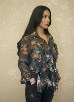 Woman wearing a dark brown floral ruffled blouse with long sleeves, styled with dark jeans, standing against a light textured wall. The blouse has soft ruffles and earthy floral patterns.