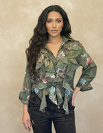 Woman in a green floral ruffled blouse with tie-front detail, paired with distressed jeans. The blouse has layered ruffles and a botanical print, creating a soft feminine look.
