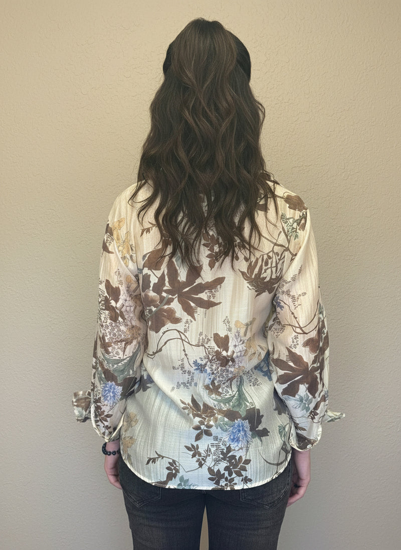Back view of woman wearing a cream and brown floral blouse with long sleeves and soft pleats, paired with dark jeans.