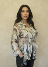Woman wearing a cream floral ruffled blouse with brown and blue tones, styled with dark jeans, standing against a beige textured wall.