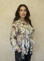 Woman wearing a cream floral ruffled blouse with brown and blue tones, styled with dark jeans, standing against a beige textured wall.