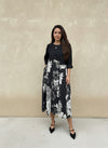 Model in a chic black and white floral midi dress with a solid black top and pleated floral skirt, posing against a neutral beige wall in black pointed-toe heels