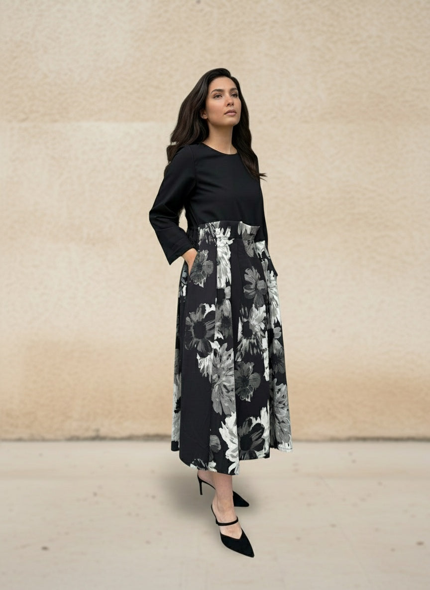 Woman wearing an elegant black midi dress with long sleeves and a monochrome floral skirt, standing sideways against a beige textured wall. Stylish modern look with black heels