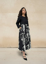 Woman wearing an elegant black midi dress with long sleeves and a monochrome floral skirt, standing sideways against a beige textured wall. Stylish modern look with black heels