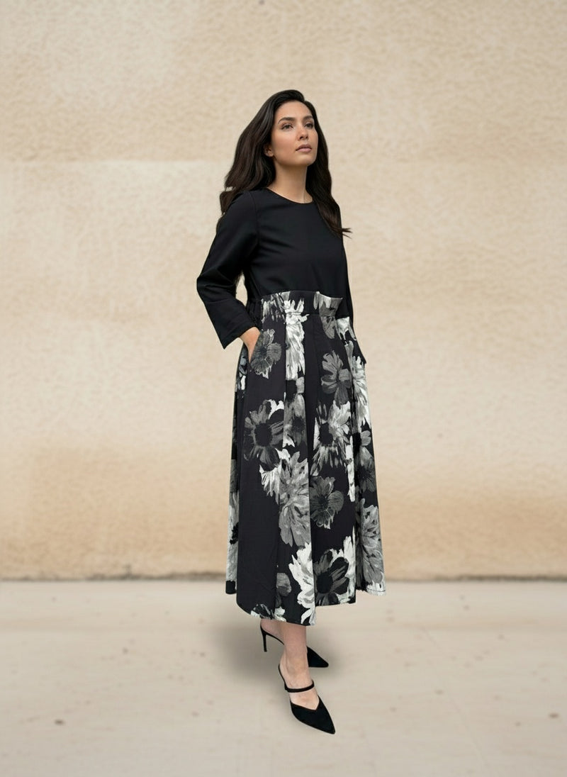 Woman wearing an elegant black midi dress with long sleeves and a monochrome floral skirt, standing sideways against a beige textured wall. Stylish modern look with black heels