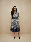Woman wearing a black and white floral print midi dress with a grey belt, standing against a neutral beige wall — elegant fall outfit.