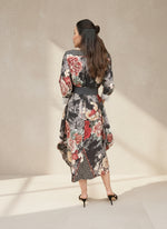 Back view of a woman wearing a black floral balloon-hem dress with large red and beige rose patterns and a dark belt, standing in a softly lit interior setting.