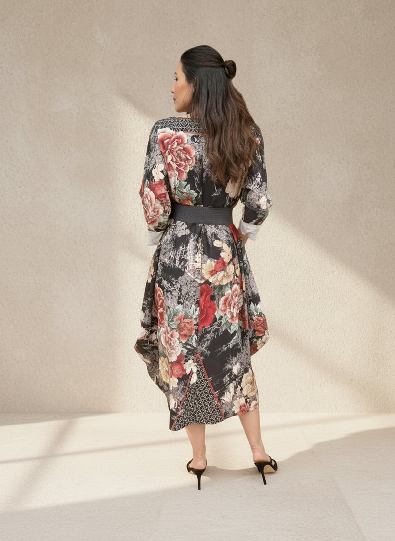Back view of a woman wearing a black floral balloon-hem dress with large red and beige rose patterns and a dark belt, standing in a softly lit interior setting.