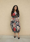 Woman wearing a black floral midi dress with red, beige, and green rose prints, cinched at the waist with a wide gray belt, styled with black pointed-toe heels against a beige wall.