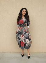 Woman wearing a black floral midi dress with red, beige, and green rose prints, cinched at the waist with a wide gray belt, styled with black pointed-toe heels against a beige wall.