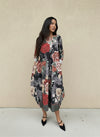 Woman in a loose black midi dress with oversized red and blush floral prints, cuffed sleeves, and patterned hem, styled with black pointed pumps against a neutral wall.