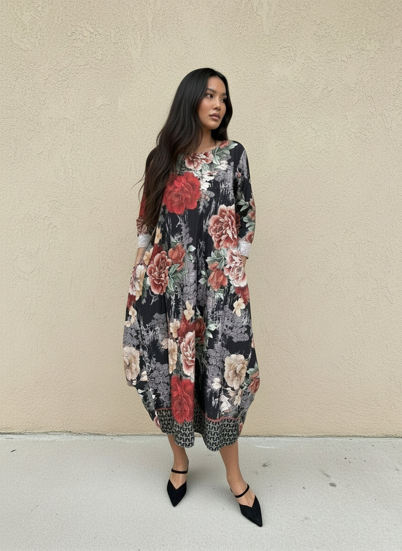 Woman in a loose black midi dress with oversized red and blush floral prints, cuffed sleeves, and patterned hem, styled with black pointed pumps against a neutral wall.