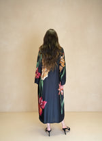 Back view of a woman wearing a black satin floral midi dress with large botanical print and long wavy hair, standing against beige background.