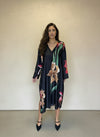 Stylish woman in black floral satin V-neck dress with long sleeves and pointed heels, posing front-facing against a neutral wall.