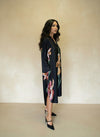 Elegant woman in a black satin floral midi dress with long sleeves, side slit, and pointed heels — side profile against beige wall.