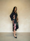Side view of woman wearing a black satin floral dress with oversized flower print and slit hem, paired with black heels.