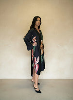 Side view of woman wearing a black satin floral dress with oversized flower print and slit hem, paired with black heels.